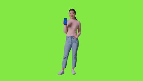 Full-Length-Studio-Portrait-Of-Smiling-Woman-Holding-Blue-Screen-Mobile-Phone-Towards-Camera-Against-Green-Screen-1